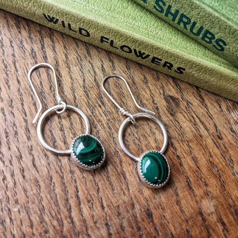A pair of earrings on a wooden surface next to some green hardback books. The earrings are small oval malachite stones mounted in a silver setting on a fixed silver hoop, on ear wires.