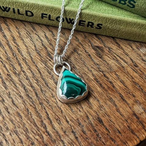 Teardrop shaped malachite pendant, set in silver on a silver chain. It is resting on a wooden surface next to some green hardback books.