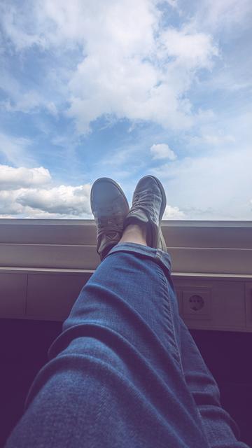 Das Bild zeigt ein Paar Beine, die in blauen Jeans gekleidet sind und entspannt auf einer Fensterbank ruhen. Die Person trägt weiße Turnschuhe, die im Sonnenlicht glänzen. Im Hintergrund erstreckt sich ein klarer Himmel mit einigen lockeren Wolken, die eine friedliche Atmosphäre schaffen. Unter der Fensterbank befindet sich eine Steckdose, die subtil ins Bild integriert ist