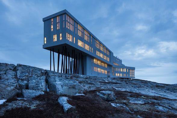 A contemporary, multi-story hotel with a flat roof and vertical windows is perched on stilts above a rugged, rocky shoreline. This is the Fogo Island Inn in Newfoundland, Canada, showcasing sustainable architecture and design.