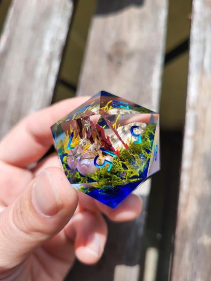 An oversized D20 with a sea-life inspired diorama inside, featuring shells, pearls, and green moss on a blue base. The numbers are inked in a colour gradient from dark blue to teal.