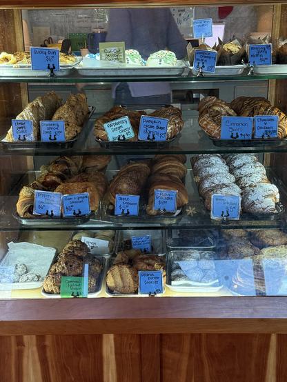 Bakery display filled with a variety of pastries, including scones, muffins, and croissants. Blue price tags describe each item. Warm, inviting ambiance.