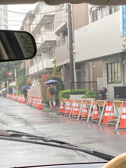 A street scene in rainy weather featuring two people walking with umbrellas. The area is lined with orange and white construction cones and reflects puddles on the ground. Apartment buildings and greenery are visible in the background.
