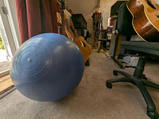 A photograph of a large blue Reebok exercise ball sitting on the floor of the White Room at Peter Gabriel's recording studios near Bath in the UK. It is taken from low level so that the ball takes up much of the frame. An open doorway and a set of orange curtains are visible at the far left. Behind the ball you can see a rack of acoustic guitars and behind that, an upright piano, a hat stand that is being used for storing cables, an Akai keyboard, and a rack of outboard processing gear. To the right, a battered acoustic guitar has been placed precariously on an office chair.