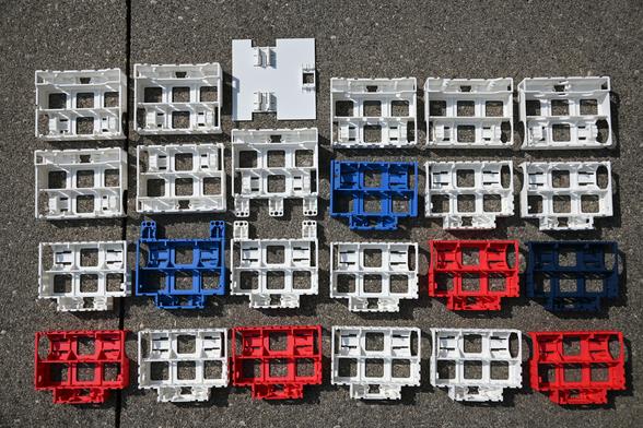 Top down view of twenty-four white, red, and blue 3D-printed iterations of the BEAPER Bot robot chassis sitting outside on grey concrete.