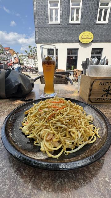 Ein Teller mit gebratenen Nudeln mit Garnelen und Gemüse steht auf einem Tisch. In der Nähe ist ein Glas Weizenbier zu sehen, zusammen mit einigen Utensilien und einer Tüte. Im Hintergrund sind ein Gebäude und Menschen in einer lebhaften Außenumgebung zu sehen.