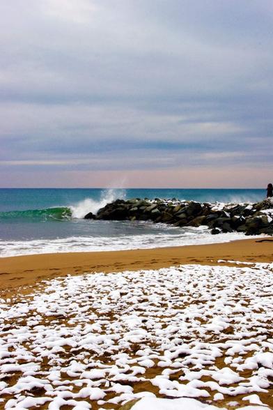 Photo au format vertical d’une plage de sable ocre et d’une digue de rochers noirs. Le ciel est couvert par une épaisse couche de nuages gris, tirant vers le rose au dessus de l’horizon. La mer a une jolie teinte bleu-vert foncé, et une vague se brise sur la digue. Au premier plan, une fine couche de neige recouvre le sable.
