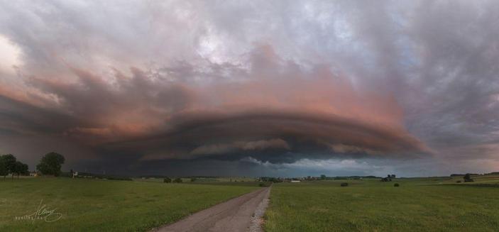 Foto einer gigantischen Gewitter Superzelle mit Ausläufern, die wie Finger in Windrichtung zu greifen scheinen. Der Kranz um das Auge des Sturms ist apokalyptisch rot angestrahlt von der untergehenden Sonne. Darunter bricht sich strahlend blau etwas schöner Himmel von der anderen Seite.