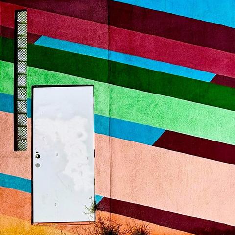 Plain white door on a building painted in bright diagonal lines of varying colors, including greens, reds, oranges, and blues. To the left of the door is a narrow vertical glass block window that is only one glass block wide but ten blocks tall.