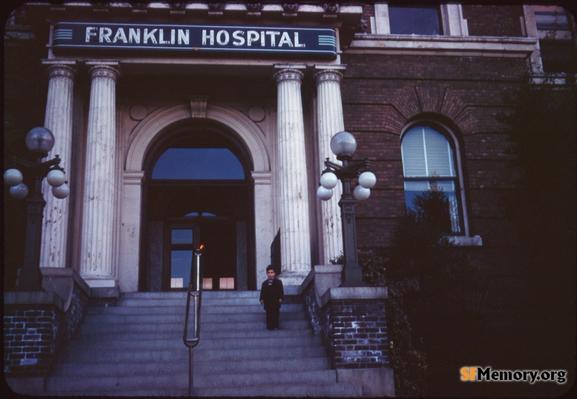 boy posed on the steps of Franklin Hospital, on the hill between Noe & Castro, 14th & Duboce. White Columns, steps, and a neon sign reading "Franklin Hospital"