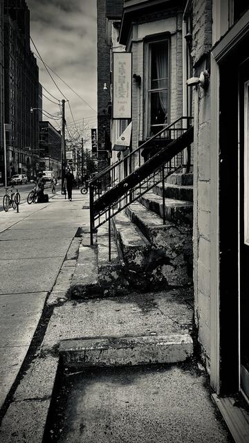 Black and white streetscape shows side view of a staircase, lots of uneven concrete and doorways and a bit of a bay window at an oblique angle. The effect is weathered and decaying.