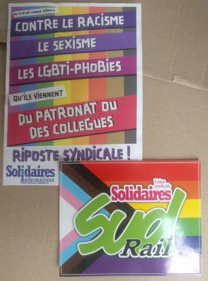2 stickers posé sur un carton :

Sur le premier est écrit "Au travail comme ailleurs, contre le racisme, le sexisme, les LGBTI-phobies qu'ils viennent du patronat ou des collègues : Riposte Syndicale !" sur fond de drapeau arc-en-ciel

Sur le second il y a le logo de Sud Rail avec un fond de drapeau arc-en-ciel avec une flèche formée par deux bandes noire et brune puis 3 bandes bleue rose et blanche.
