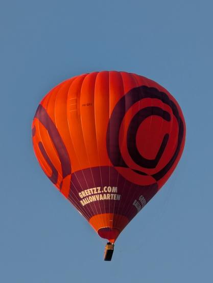 A vibrant orange and purple hot air balloon, bearing the 'GREETZZ.COM BALLONVAARTEN' logo, ascends against a clear blue sky. The balloon's upper section is orange with large, dark purple swirling patterns, while the lower part is predominantly purple with the website address in white text. A basket is visible beneath the balloon. The registration code 'PH-GRU' is faintly visible on the envelope.