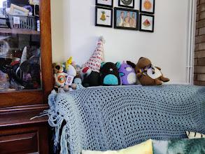 A photo of stuffed animals lined up on the back of the radiator, behind a couch covered in a blue crochet blanket. From left to right, there is a small meerkat dressed up as the bot BB-8 from Star wars, a hand-knit unicorn, a hand-knint monkey, a garden gnome plus with a 'ho ho ho' print hat, a black and blue Squishmallow bat, a purple and white Squishmallow unicorn, and Pudding the Reindeer, who is round and wearing a striped scarf.