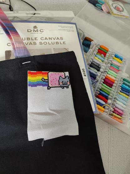 Eine schwarze Stofftasche mit bunten Trägern, darauf liegt eine auf grauem Aida gestickte Nyancat, neben wasserlöslichen Zählstoff und einer aufgeklappten Sortierbox voller Stickgarn in allen möglichen Farben.