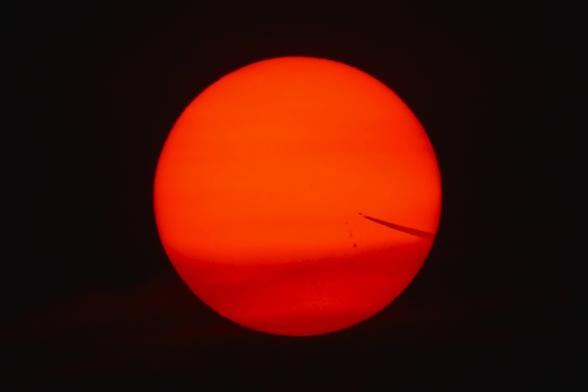 Photograph of a bright red and orange Sun with its light filtered through smoke and ash from the Canadian wildfires. A jet and contrail are visible headed towards the center from the right side. Sunspots are visible below the jet.