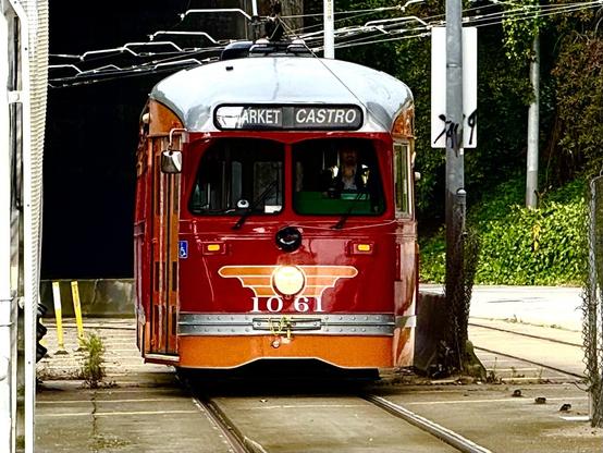 A vintage red and orange streetcar is emerging from a tunnel onto a set of streetcar tracks. The streetcar has a rounded front with the route sign reading “MARKET CASTRO” above the windshield. The vehicle number “1061” is prominently displayed on the front, below a stylized winged emblem. The streetcar is equipped with overhead trolley poles connected to electric wires above.