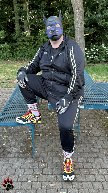 human pup wearing a Nike tracksuit, Nike football gloves, white socks with text 'FCK ABL' visible and Nike Shox sneakers half-sitting on a table at a German motorway rest area, looking into the camera