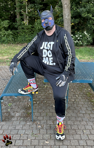 human pup wearing a Nike tracksuit, Nike football gloves, white socks with text 'FCK ABL' visible and Nike Shox sneakers half-sitting on a table at a German motorway rest area, looking into the camera, tracksuit top is opened to reveal a t-shirt with text 'just do me' on it
