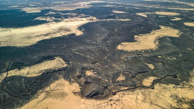 An oblique aerial view of an ancient animal hunting trap called a "Desert Kite". The main part of the trap is in the foreground, a stone enclosure with small circular "cells" spaced around its circumference. In the distance the walls that guide the animals into the trap can be seen stretching out into the distance.