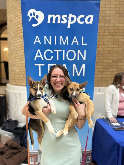 an mspca advocacy staffer in a mint green dress holds two shepherd mix puppies and smiles broadly. she's standing in front of a blue mspca animal action team banner. she has long brown hair and glasses