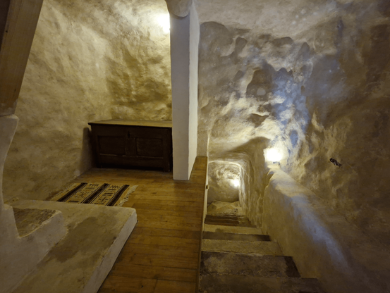 Les marches pour accéder au niveau supérieur dans la maison troglodyte, creusées dans une espèce de tunnel aux parois arrondies aux surfaces blanches. Dans un coin, sur le palier entre les étages, un petit meuble noir.