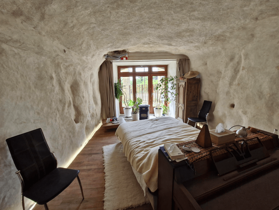 Une chambre avec un grand lit dans une grotte aux parois blanches, avec une baie vitrée pour faire entrer la lumière et des plantes pour encadrer les fenêtres.
