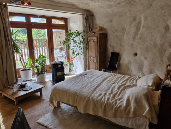 Une chambre avec un grand lit dans une grotte aux parois blanches, avec une baie vitrée pour faire entrer la lumière et des plantes pour encadrer les fenêtres.
