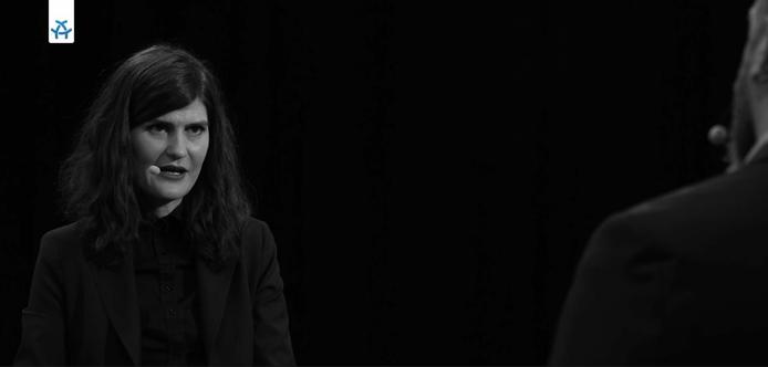 Schwarz-weißes Still aus dem Interview. Rechts die Schulter des Hosts, links weiter hinten sitze ich mit einem Ansteckmikrofon vor dunklem Hintergrund.
