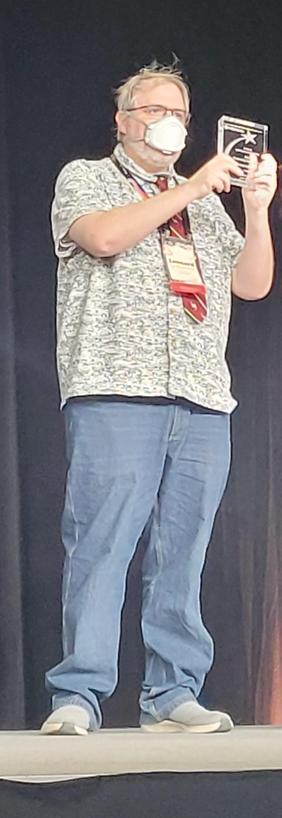 Leonard on a stage, holding up a rectangular glass object (the award)