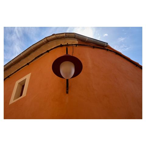 Image of a building with a curved, ochre-coloured wall. The camera is pointed upwards. On the left, a window with a white frame is visible. In the center, a street lamp with a circular cover is attached to the wall. The top of the frame has a bit of blue sky.