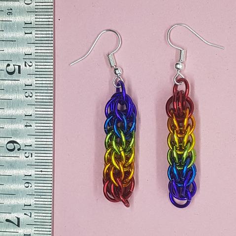 A pair of earrings made of chainmail, against a pink backdrop.
Rainbow colours. Left has purple at the top, red at the base. Right has red top, purple base.
There is a steel ruler on the left to show their size of approx 1.5 inch