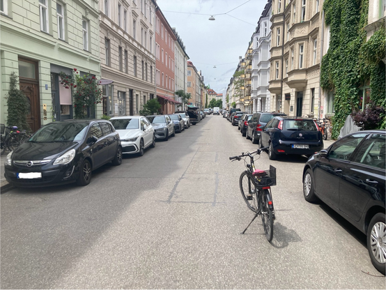 Straße in München. An den Fahrbahnrändern parken Autos. Einen Meter neben dem Auto steht ein Fahrrad. Rechnet man den Sicherheitsabstand von 1,5 Meter noch dazu, dann bleibt kein Platz mehr für ein fahrendes Auto, um zu überholen.
