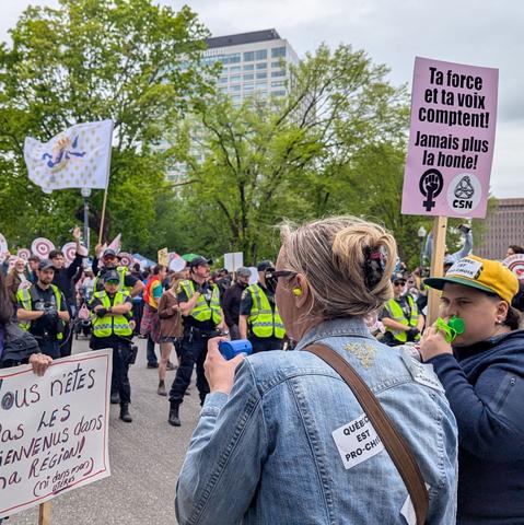 Des manifestantes tiennent des affiches "ta force et ta voix comptent! Plus jamais la honte!" et "Vous n'êtes pas les bienvenus dans la région! (ni dans mon utérus) .  L'une d'entre elle souffle dans une espèce de sifflet vert. Il y a plusieurs autres manifestants , devancés par des policiers aux vestes jaunes fluorescentes.  Le ciel est gris, en arrière-plan, de grands arbres feuillus cachent un édifice gris en hauteur, le Hilton.