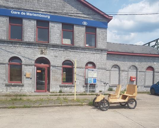 à l'arrière plan le bâtiment girs aux fenêtre avec arcades de la Gare de Mariembourg (on y vois le bandeau bleu avec le signe B de la sncb et le texte "gare de Mariembourg)
devant, un quai de bus sur la place avec quelques herbes sauvages et le Shifti, quadricycle partiellement en bois, garé sur la droite de l'image