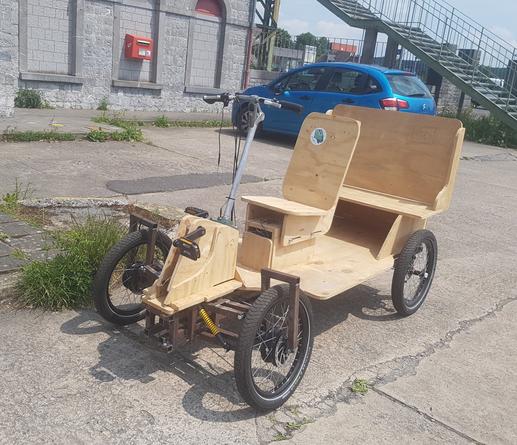 une vue de trois quart du véhicule léger montrant plus distinctement ses sièges an bois, une place conducteur et banquette arrière sur son chassi en métal flanqué de quatres roues de vélo. on distingue les moteurs sur les roues avant, un pédalier pour le conduteurice.