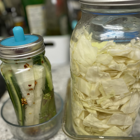 A jar with cucumber, daikon, and asparagus in brine, beside a 2-litre jar of fresh sauerkraut