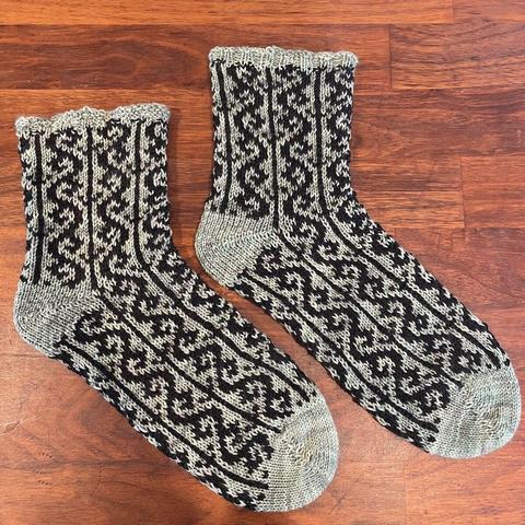 Two hand knit socks are lying parallel on a wooden surface. They have solid sage green toes, heels, and rib edge, with an intricate pattern in sage green and black yarns throughout the foot and ankle.