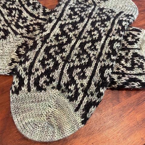 Detail of the toe of a hand knit sock. The toe is sage green, with the ankle foot of the sock knit in an intricate pattern in sage green and black yarns.