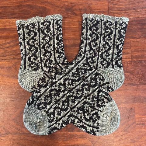 Two hand knit socks are lying crossed over each other on a wooden surface. They have solid sage green toes, heels, and rib edge, with an intricate pattern in sage green and black yarns throughout the foot and ankle.