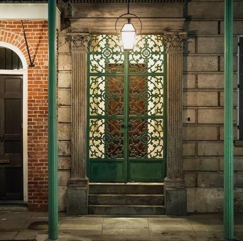 A photo of a doorway in the French Quarter of New Orleans at night, where a recessed door is partially concealed by a sage green curling wrought iron screen.