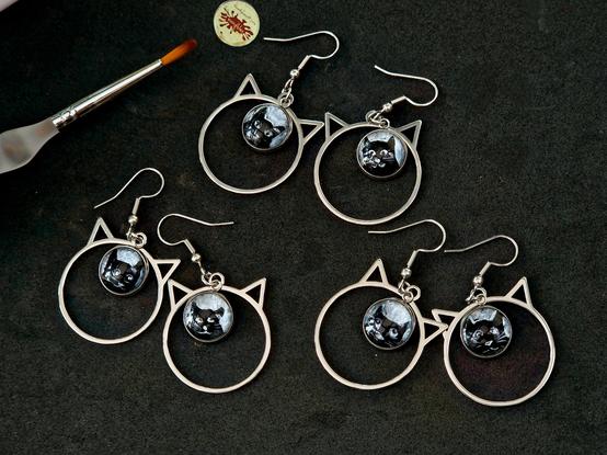 Ohrringe aus Edelstahl, haben die Form von Katzenkopf-Umrissen, und eingesetzte baumelnde Anhänger mit handgemalten 12mm-Motiven,mit stilisertem schwarzem Katzenkopf im ulkigen Stil