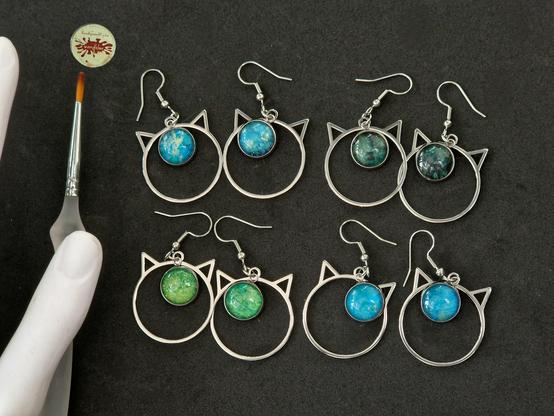 Ohrringe aus Edelstahl, haben die Form von Katzenkopf-Umrissen, und eingesetzte baumelnde Anhänger mit handgemalten 12mm-Motiven,mit farbigen Galaxien in 4 verschiedenen Grün- und Blautönen