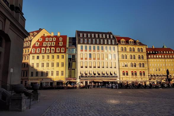A wide shot of Dresden's Neumarkt square in the warm glow of late afternoon sun. A row of beautifully reconstructed Baroque buildings, including the Aparthotel Altes Dresden, with their distinctive red-tiled roofs and pastel-coloured facades, line the square. The square itself is paved with cobblestones, and several outdoor seating areas for restaurants and cafes are visible with people enjoying the evening. The clear blue sky provides a striking contrast to the illuminated buildings.