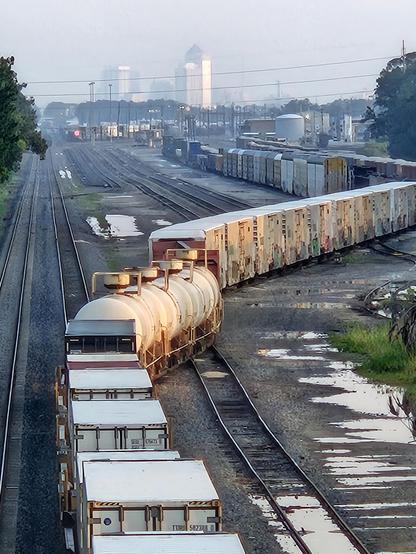 Long train of box cars winds onto tracks leading from a rail yard with the city skyline in the background.