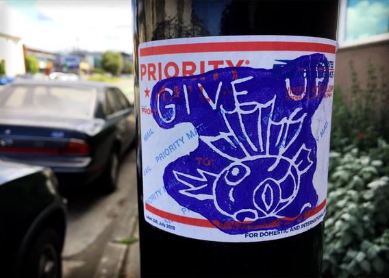 A USPS label stuck on a post. There is art on the label showing a fish laying on it's side and text that says "give up". This art appears to have been made on a woodblock then printed onto the sticker in purple ink.