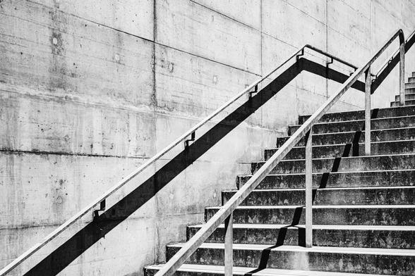 Black and white photo of a concrete outdoor staircase casting sharp angular shadows on the wall beside it. The metal handrails create bold diagonal lines, emphasizing the minimalist, geometric architecture.