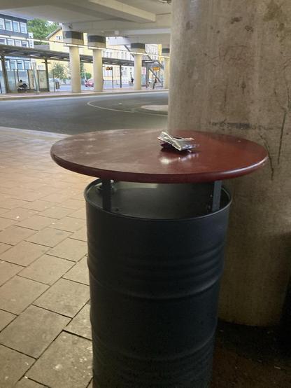 Zu sehen ist eine schwarze Metalltonne auf die mit rohrförmigen Abstandshaltern eine Tischplatte aus Holz, dunkelrot lackiert, montiert ist. Darauf liegt eine zerknüllte Caprisun. Im Hintergrund ist der Busbahnhof von Innen zu sehen.