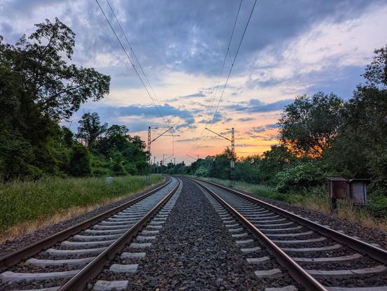 A low-angle, eye-level shot captures a railway track at sunset, stretching into the distance under a dramatic, cloudy sky. Two parallel tracks, set on a bed of gravel and concrete sleepers, curve gently to the right as they recede. Overhead lines are visible, supported by poles that are largely obscured by lush green foliage bordering both sides of the tracks. The setting sun casts a warm, orange glow on the horizon, contrasting with the cooler blue and grey tones of the clouds above. The surrounding vegetation is dense and verdant, indicating a rural or semi-rural setting.