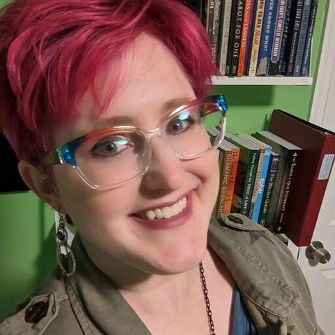 A close-up of Enfys Book, a pale-skinned person with fuchsia hair and rainbow glasses, smiling at the camera